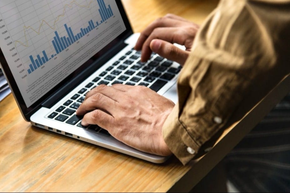 Hands typing on a laptop with a spreadsheet showing bar and line graphs on screen. The setting is a wooden desk with a focused tone.